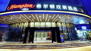 Entrada de un hotel con un llamativo letrero de 'Hampton by Hilton', fachada de vidrio moderna y un área de acceso bien iluminada.