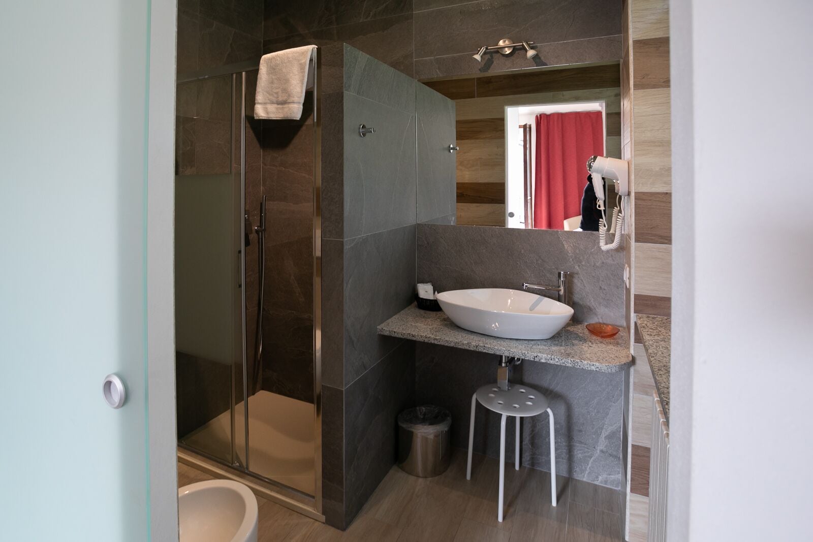 Un baño moderno con ducha de vidrio, un lavamanos blanco y un taburete pequeño.