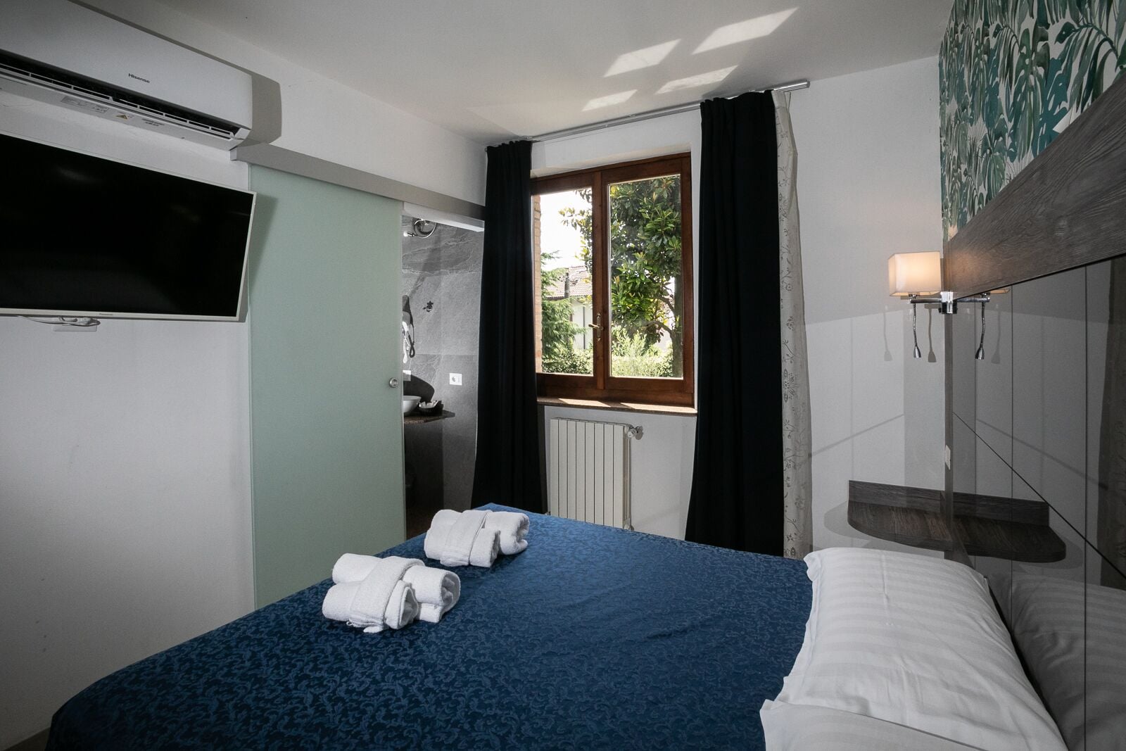 A hotel room with a bed, a TV, a window with curtains, and a wall-mounted light fixture.