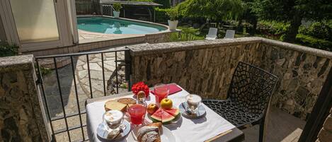 Una mesa de comedor con alimentos para el desayuno en un balcón con vistas a una piscina y un paisaje montañoso.