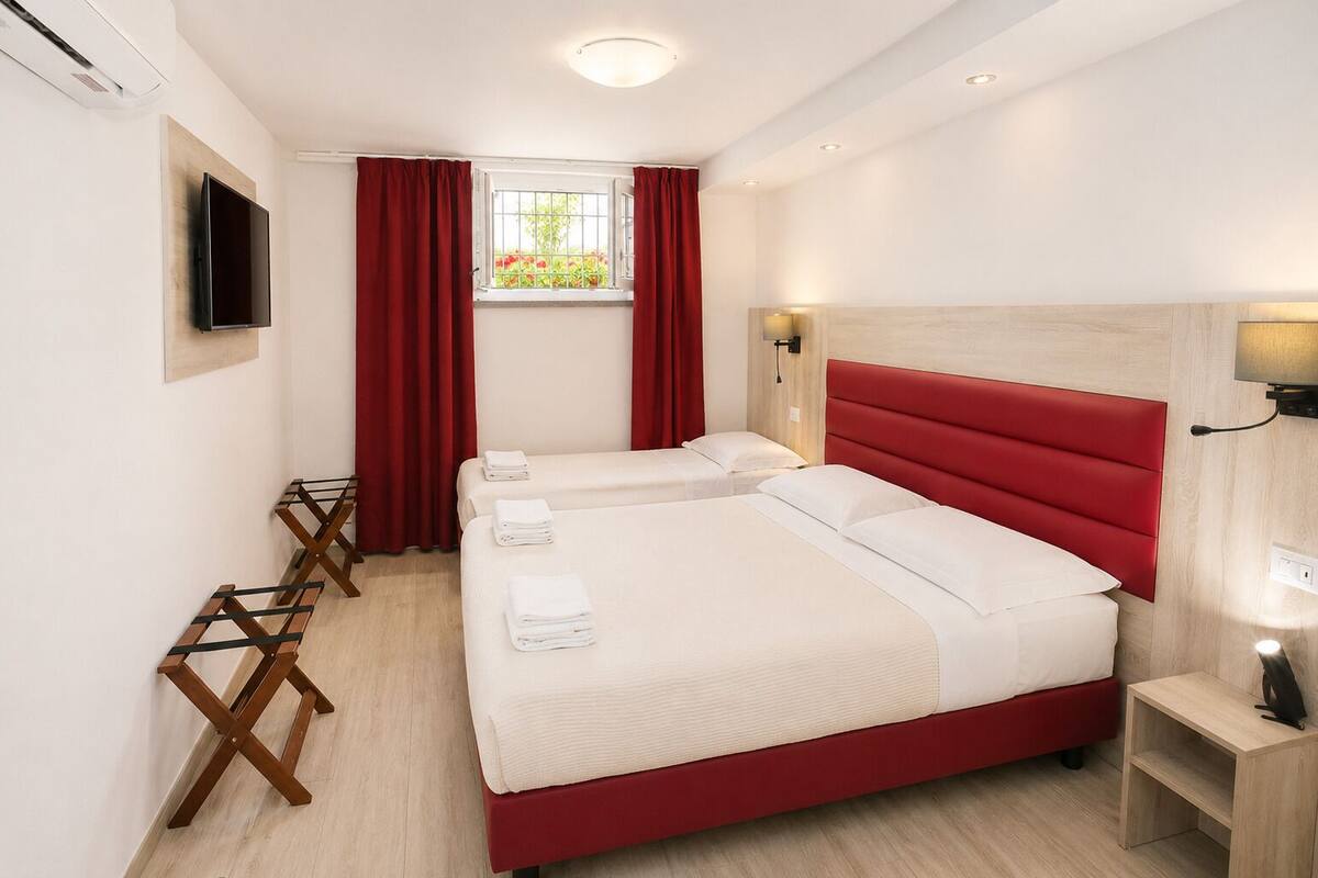 A hotel room with two beds, a TV, a window with red curtains, and a wooden floor.