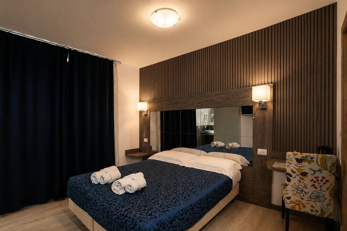 A hotel room with a large bed, a wooden headboard, a mirror, a bedside table, and a chair with a floral pattern.