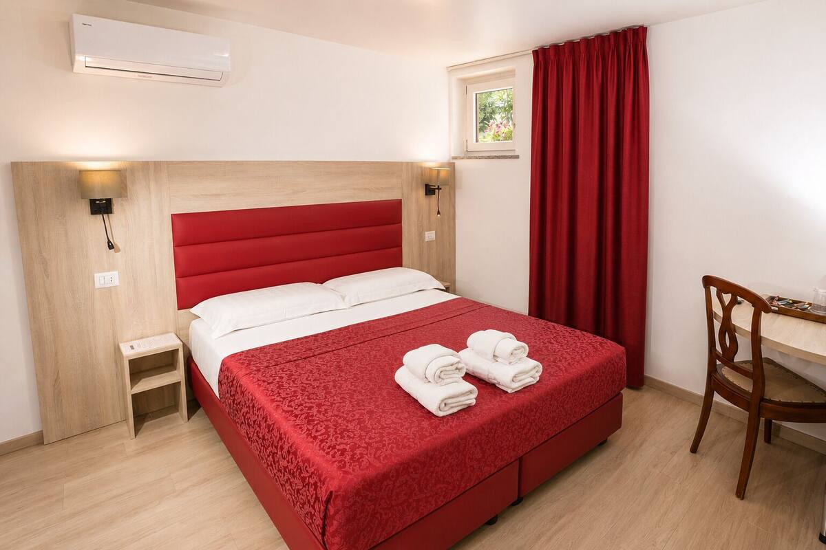 A hotel room with a red bed, a wooden nightstand, a desk with a chair, and a window with red curtains.