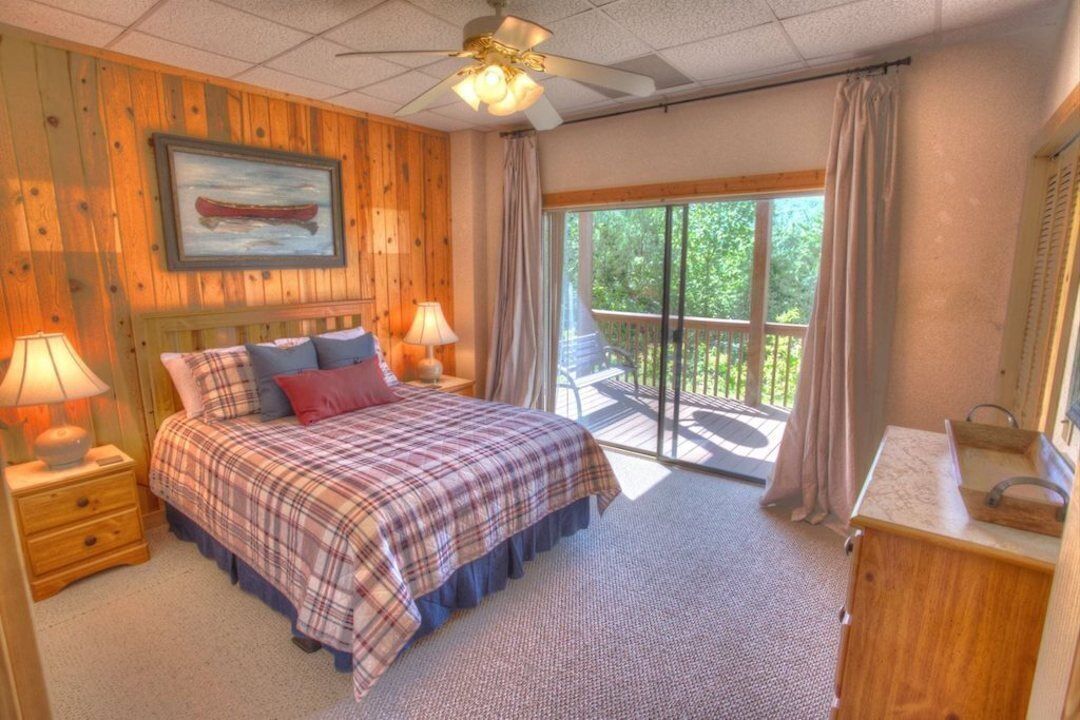 A bedroom with a bed, two bedside tables with lamps, a wooden headboard, a window with curtains, and a view of greenery outside.