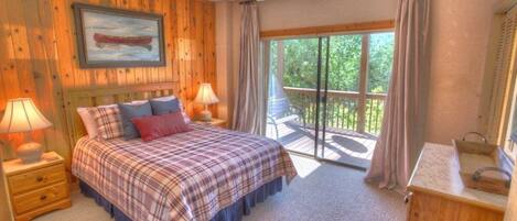 A bedroom with a bed, two bedside tables with lamps, a wooden headboard, a window with curtains, and a view of greenery outside.