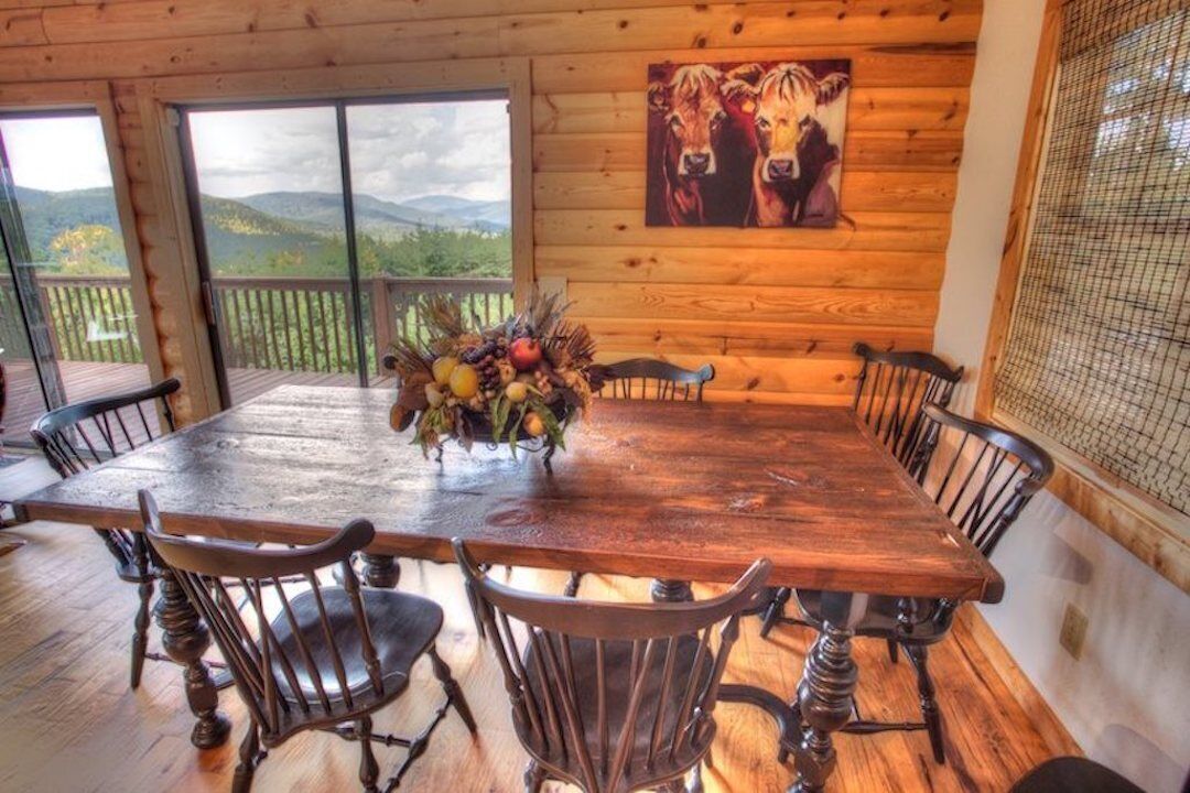 A wooden dining table with eight chairs, a centerpiece of fruit and flowers, and a painting of cows on the wall.