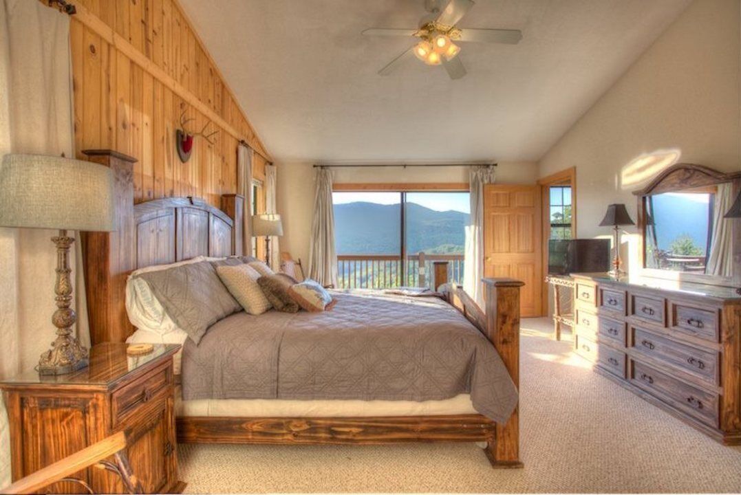 A bedroom with a large bed, wooden headboard, a nightstand, a dresser, and a balcony with a view of the mountains.