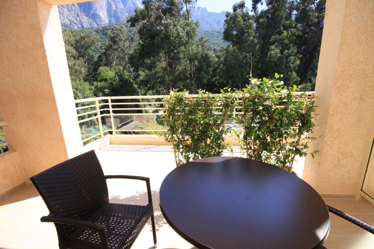 Un balcón con una silla negra y una mesa redonda, con vistas a un paisaje montañoso de exuberante vegetación.