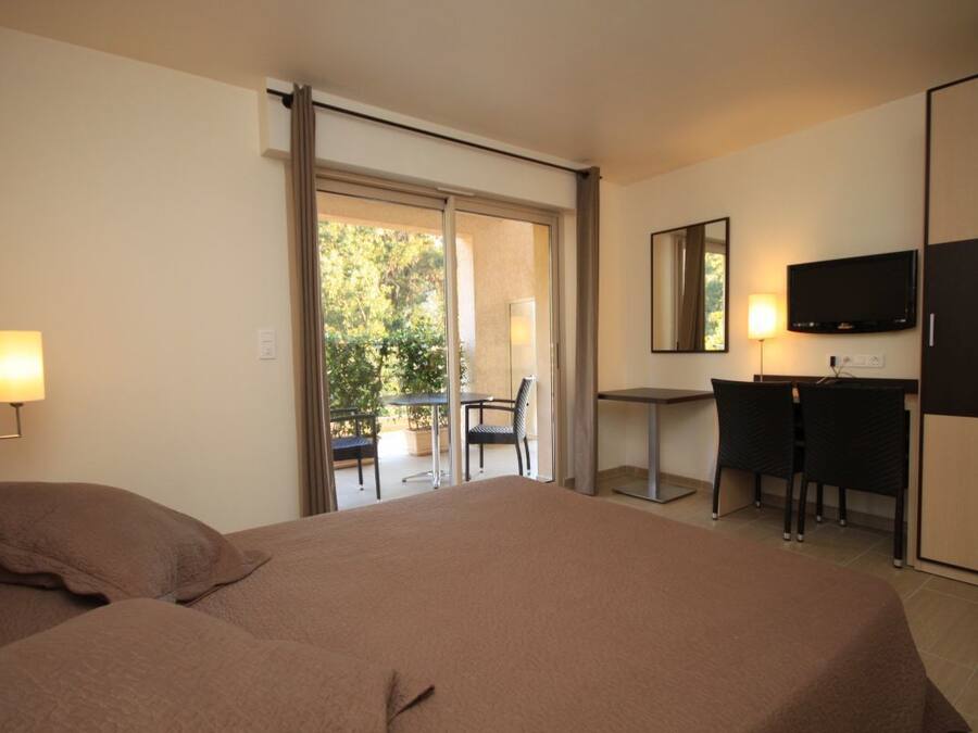 A hotel room with a large bed, a desk, a chair, a television, and a balcony with a view of trees.