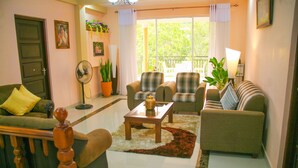 A living room with a sofa, two armchairs, a coffee table, and a fan. There is a door, a window with curtains, and a plant.