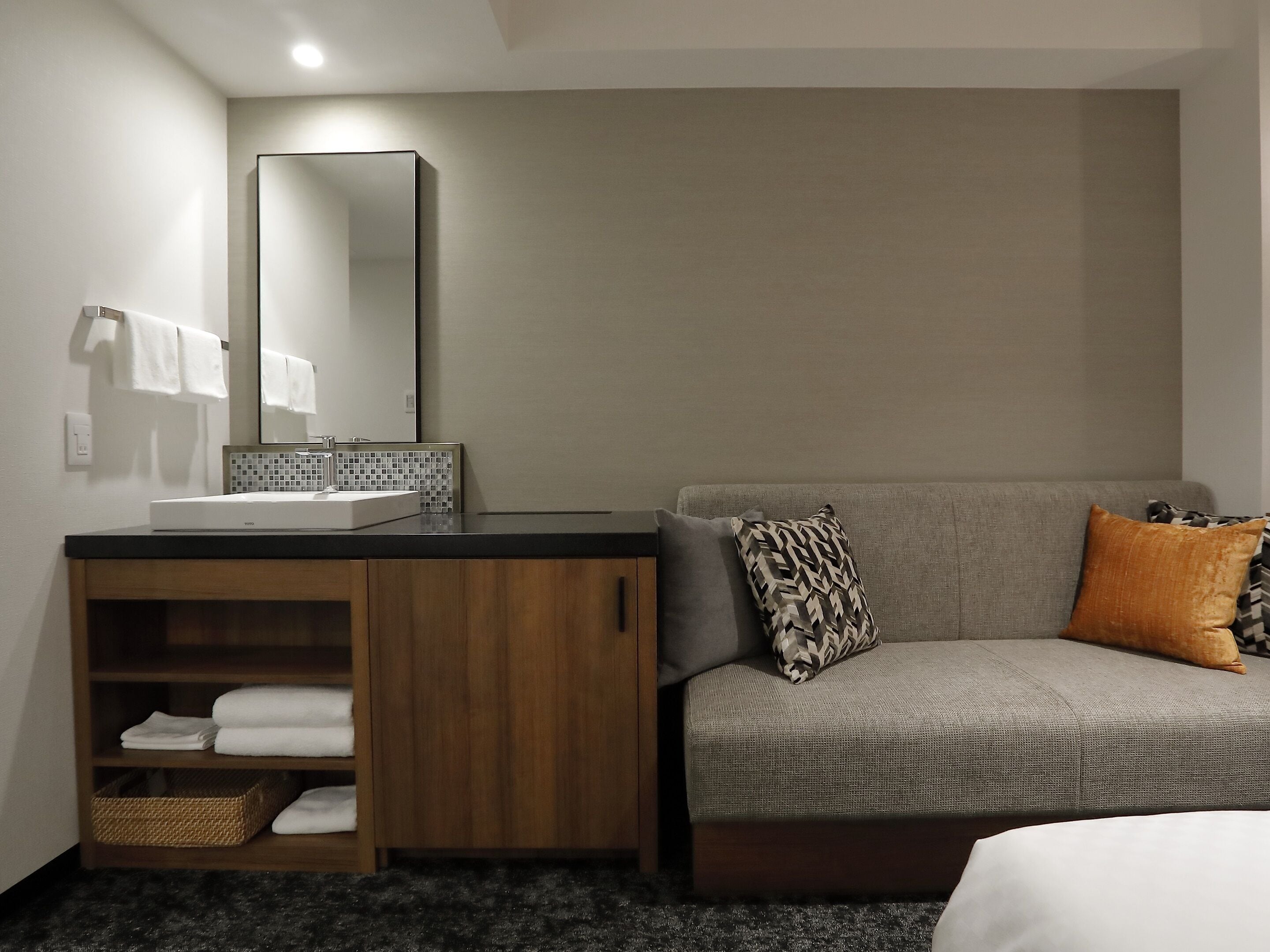 A modern bathroom with a sink, mirror, and towel rack. There is a sofa with patterned cushions and a basket of towels.