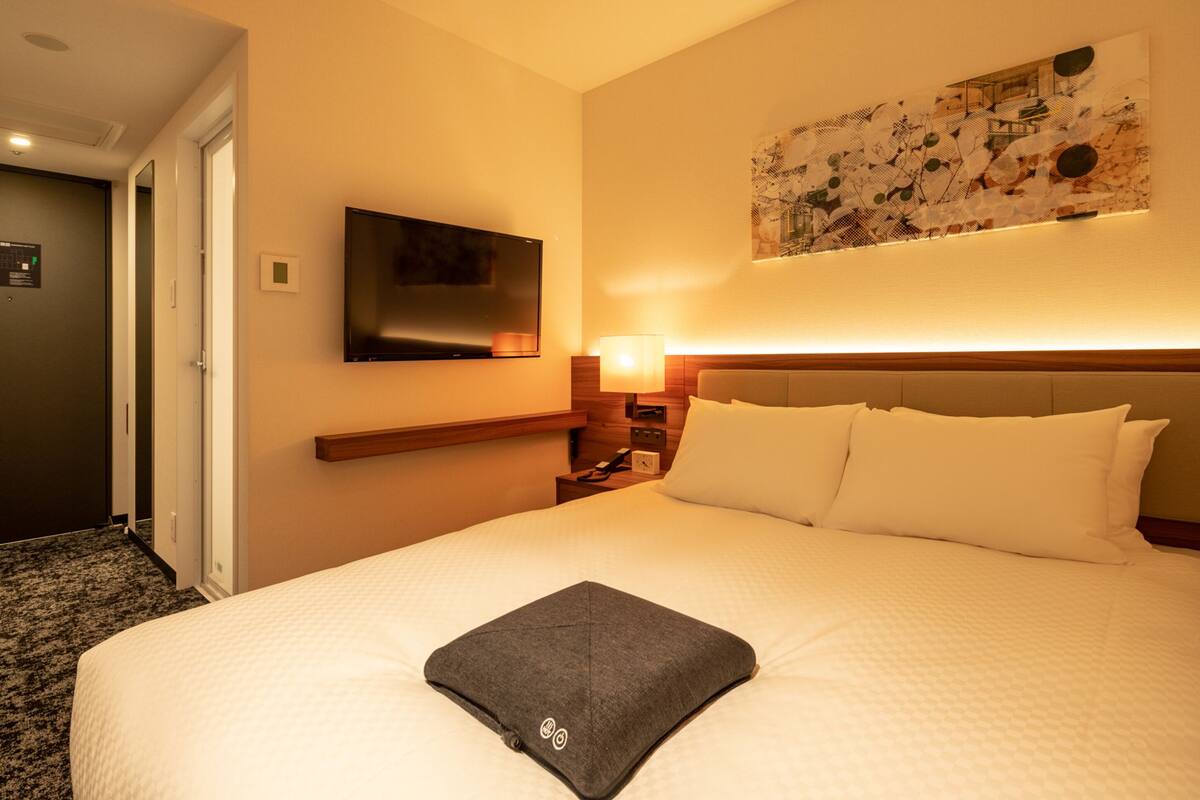 A hotel room with a large bed, a flat-screen TV mounted on the wall, a bedside table with a lamp, and a painting above the bed.