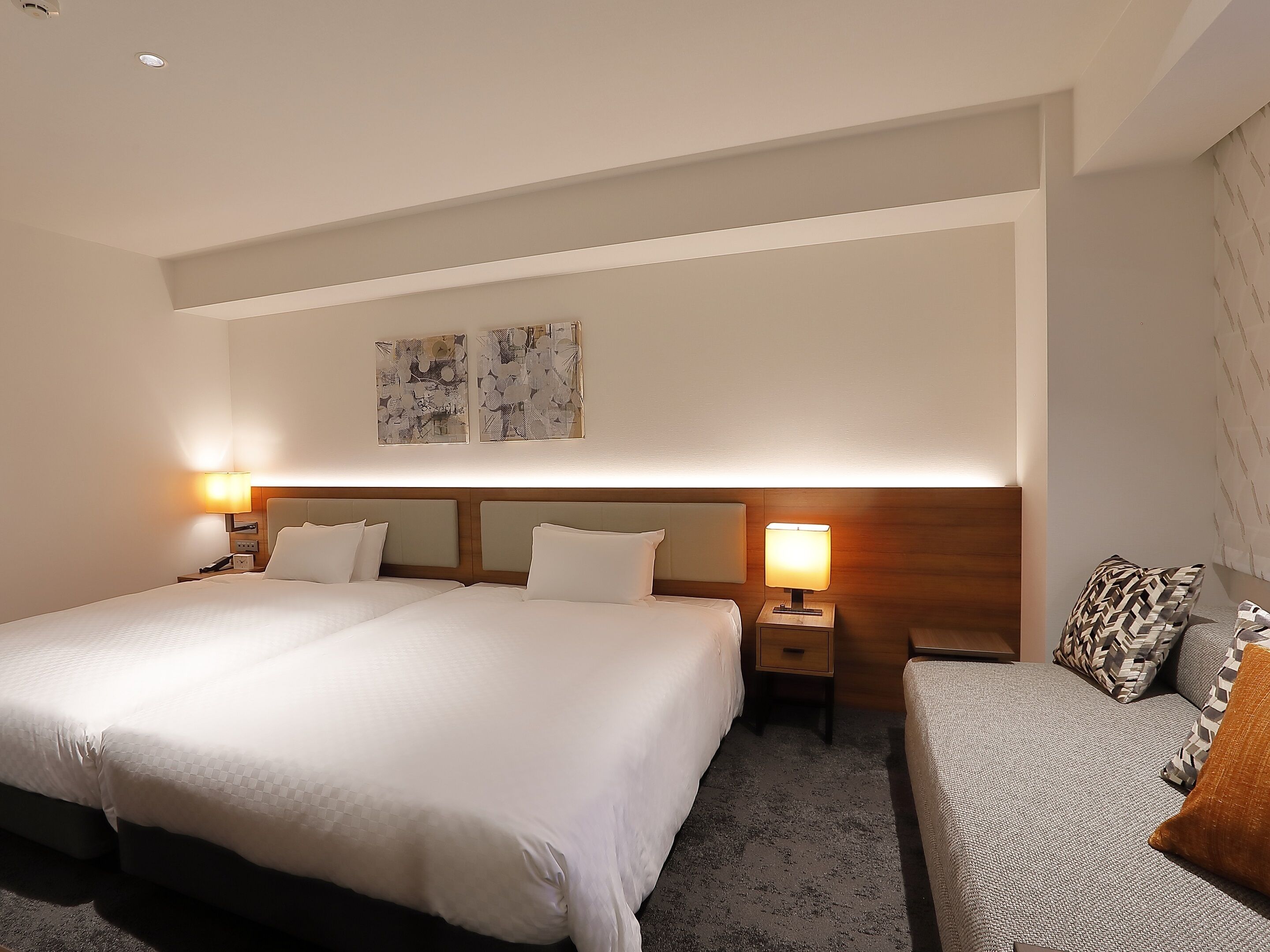 A hotel room with a large bed, a smaller sofa, two bedside lamps, and abstract artwork on the wall.
