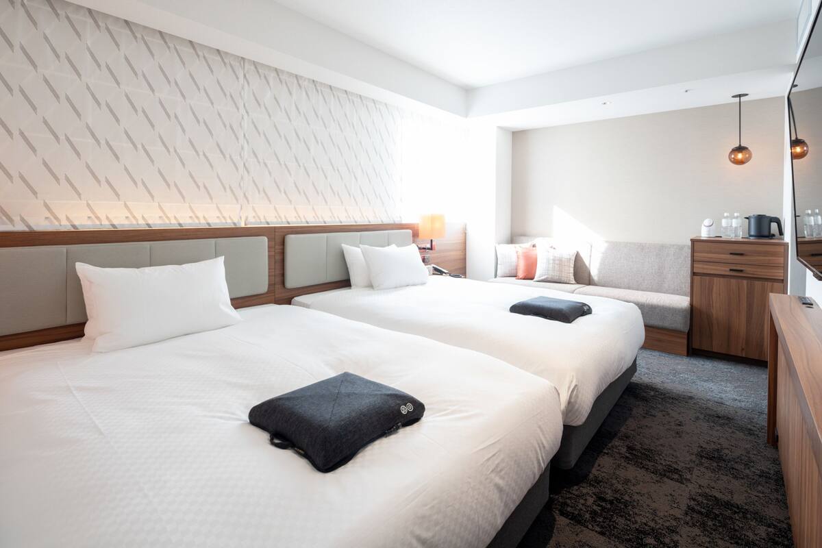 A hotel room with two beds, a wooden headboard, a nightstand with a lamp, a sofa, a wooden dresser, and a mirror.