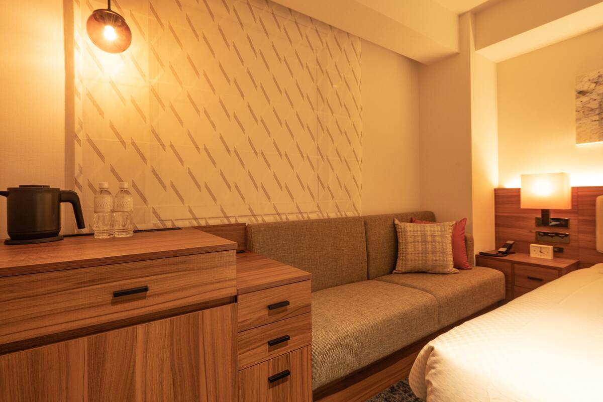 A hotel room with a wooden dresser, a sofa, a bed, and a nightstand with a lamp.