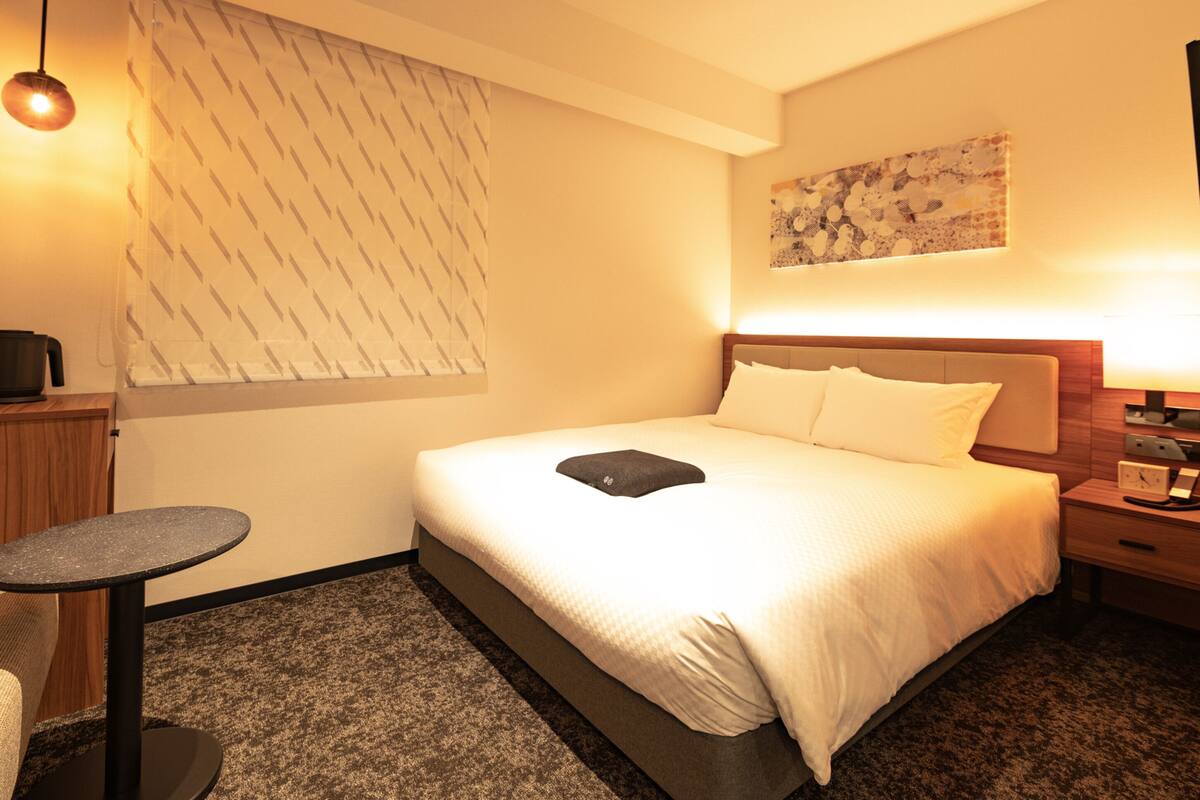 A hotel room with a bed, a small table, a nightstand, a lamp, and a wall art piece.