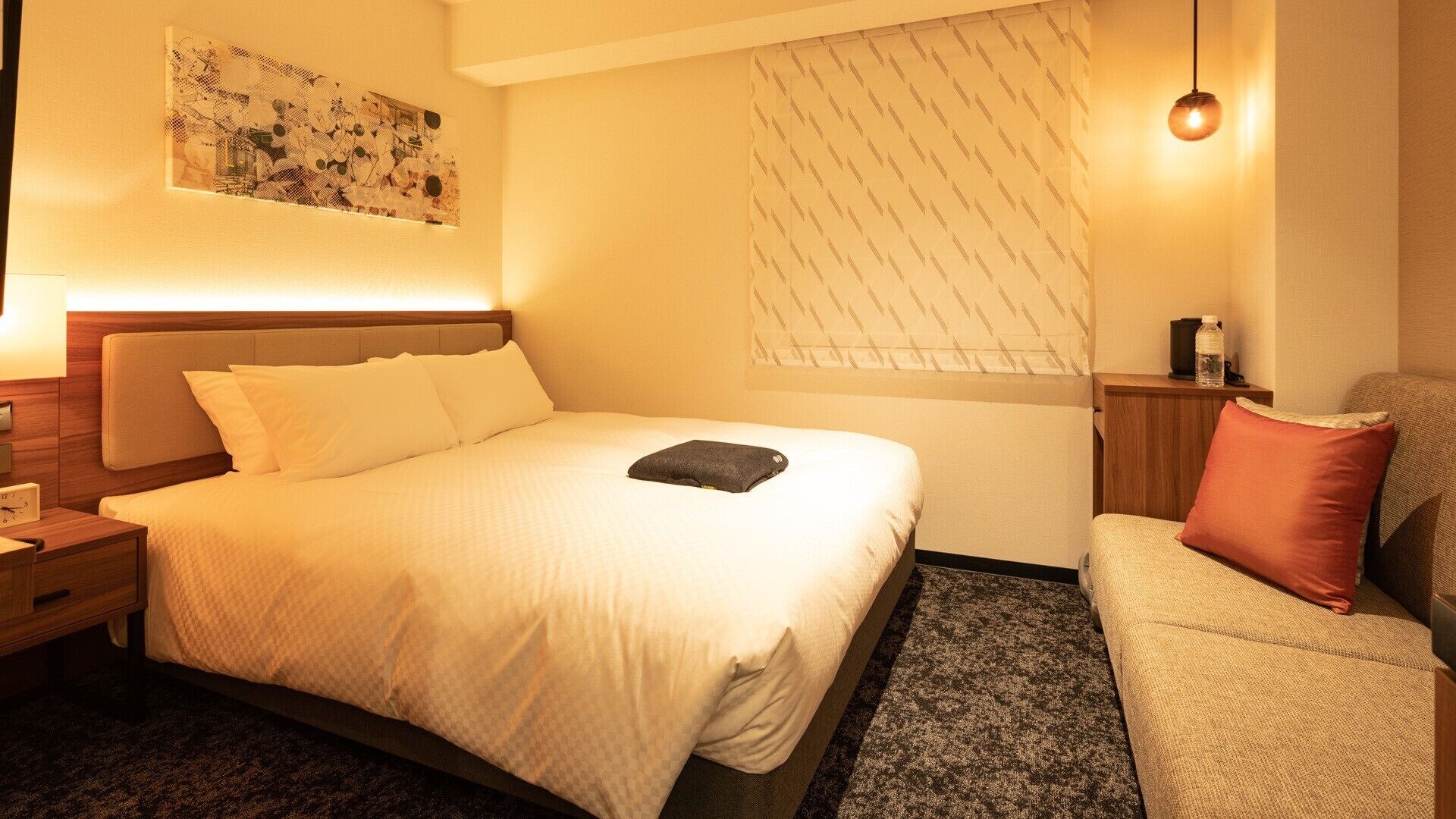 Ein Hotelzimmer mit Bett, Nachttisch, Lampe und einer kleinen Sitzecke mit Stuhl und Sofa.