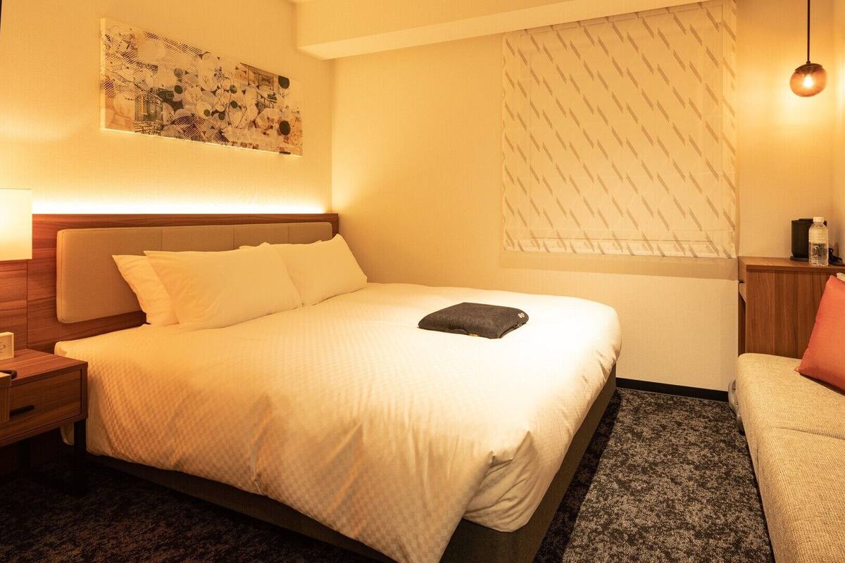 Ein Hotelzimmer mit Bett, Nachttisch, Lampe und einer kleinen Sitzecke mit Stuhl und Sofa.