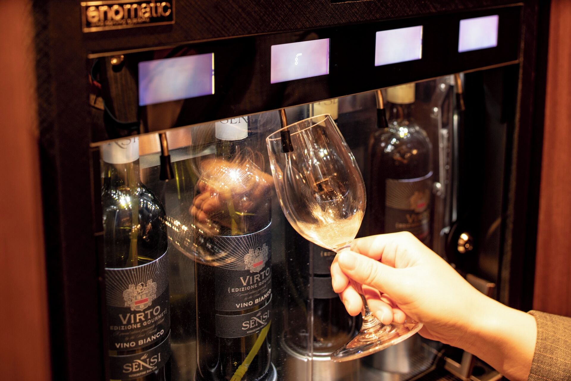 A hand holding a wine glass in front of a wine dispenser with bottles labeled 'VIRTO' and 'SENSI'.