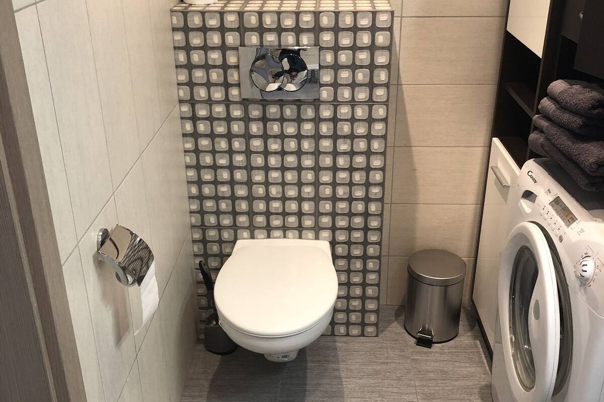 A compact bathroom with a toilet, a washing machine, and a small trash bin.