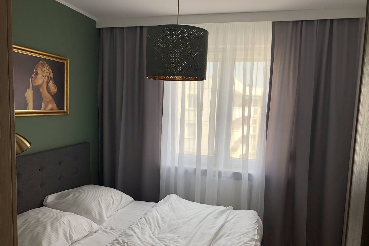 A bedroom with a bed, a hanging light fixture, a painting on the wall, and sheer curtains.