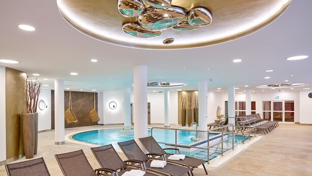 A modern indoor swimming pool area with lounge chairs and a central pool. - Maximilian Quellness- und Golfhotel (Bad Griesbach im Rottal)