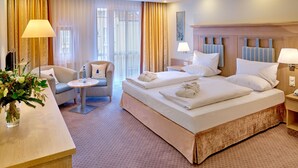 A hotel room with a large bed, a sitting area with a chair and a small table, and a desk with a lamp. - Maximilian Quellness- und Golfhotel (Bad Griesbach im Rottal)