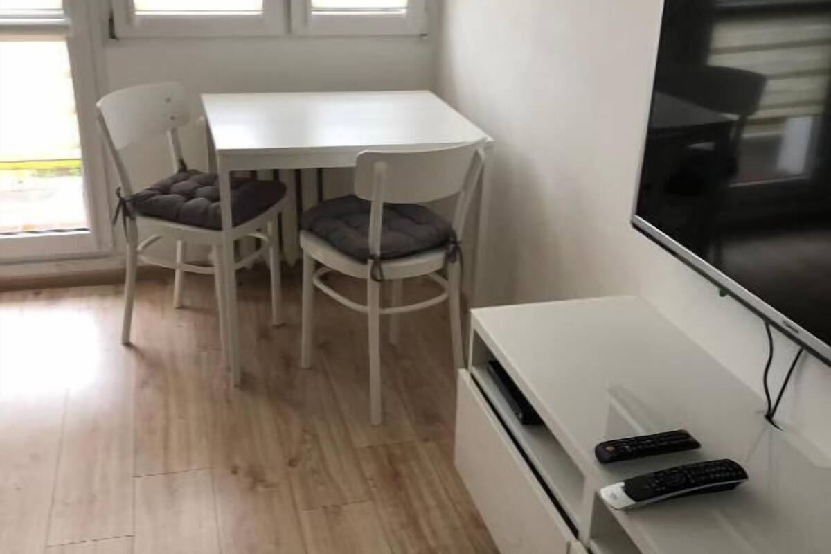 A small, bright room with a white table and two chairs, a TV on a stand, and a window with blinds.