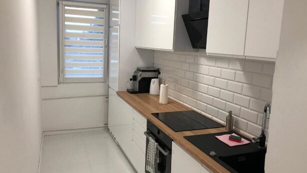 A modern kitchen with white cabinets, a black countertop, a built-in oven, and a window with blinds. - Apartament Przy Skarpie (Torun)