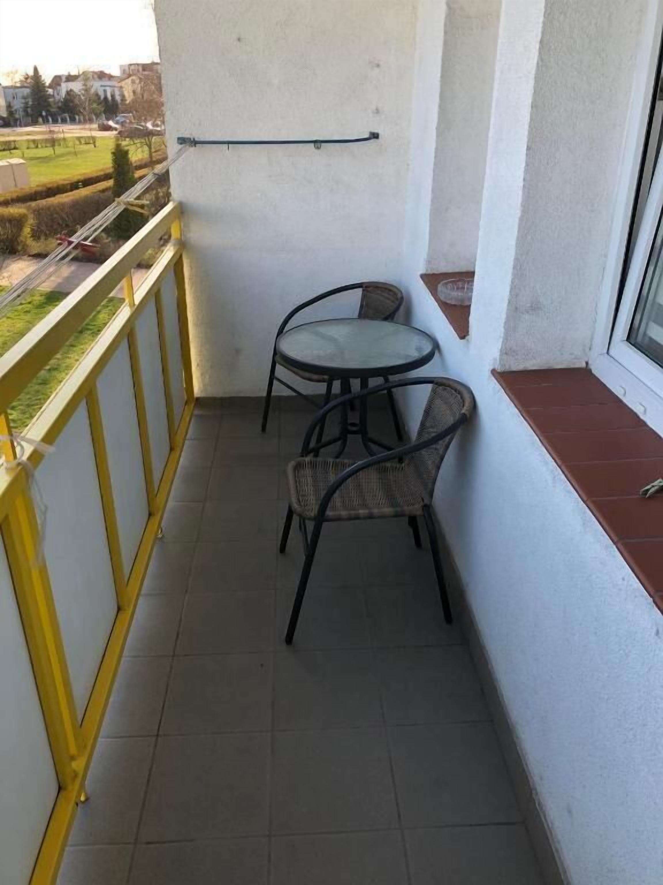 A small balcony with a table and two chairs, a window, and a view of a park.