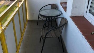 A small balcony with a table and two chairs, a window, and a view of a park.