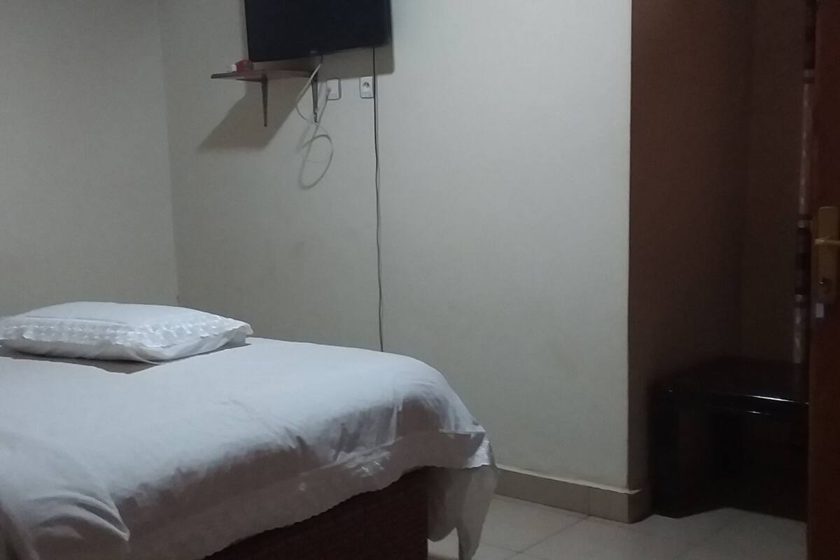 A small, dimly lit room with a single bed, a wall-mounted TV, and a door leading to another area.