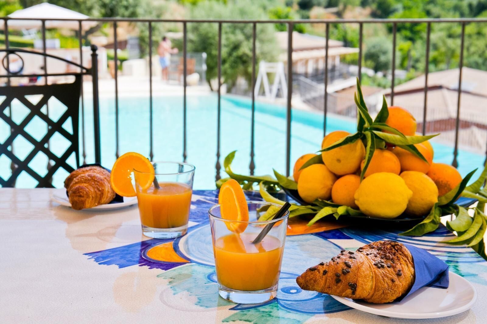 A table with a croissant, two glasses of orange juice, a plate with a croissant, and a bowl of oranges on a terrace with a pool in the background.