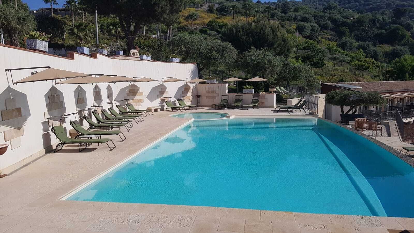 A rectangular swimming pool with a clear blue water surface, surrounded by lounge chairs and umbrellas, set against a backdrop of trees and a hillside.