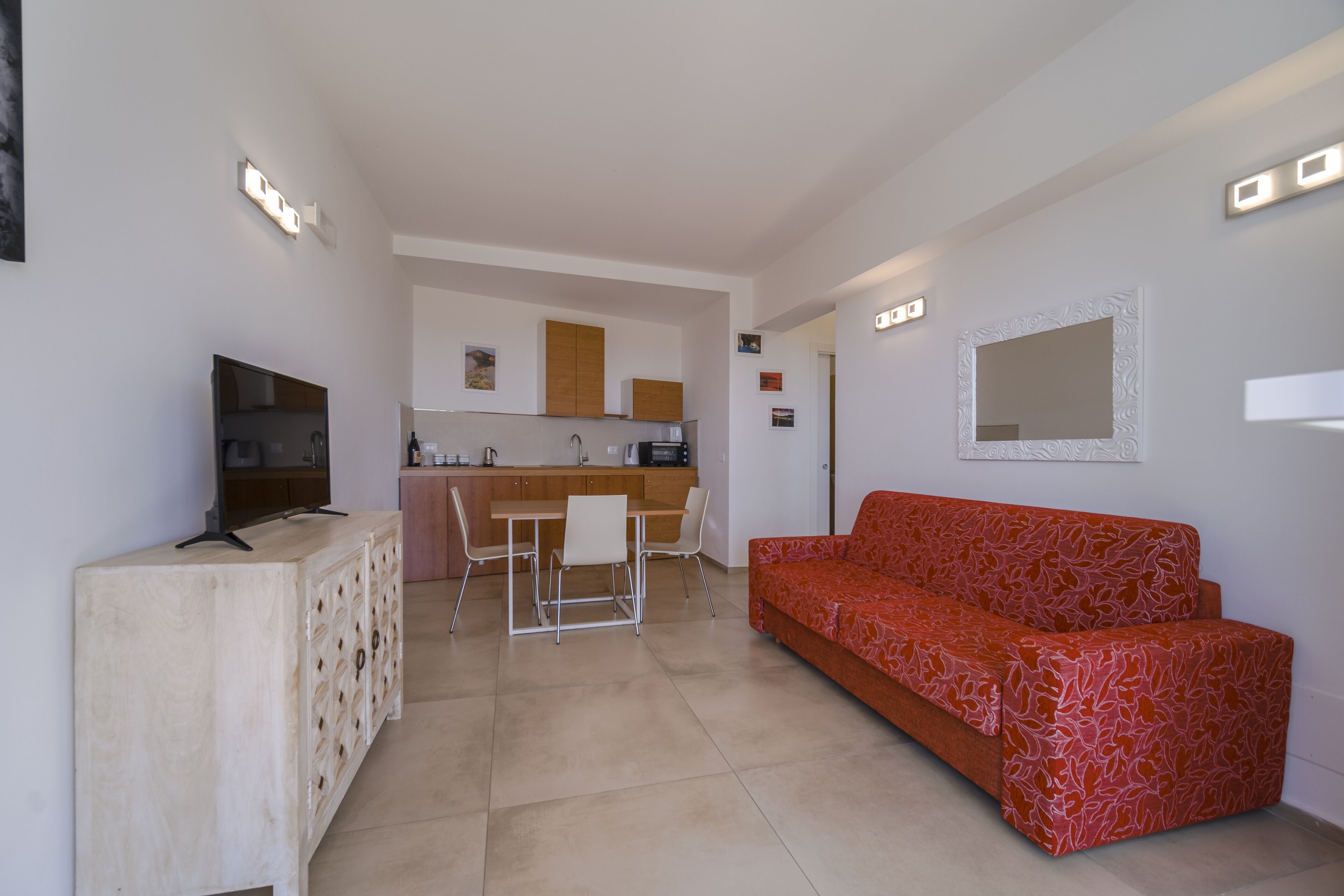 A modern living room with a red patterned sofa, a wooden cabinet with drawers, a TV, and a kitchen area in the background.