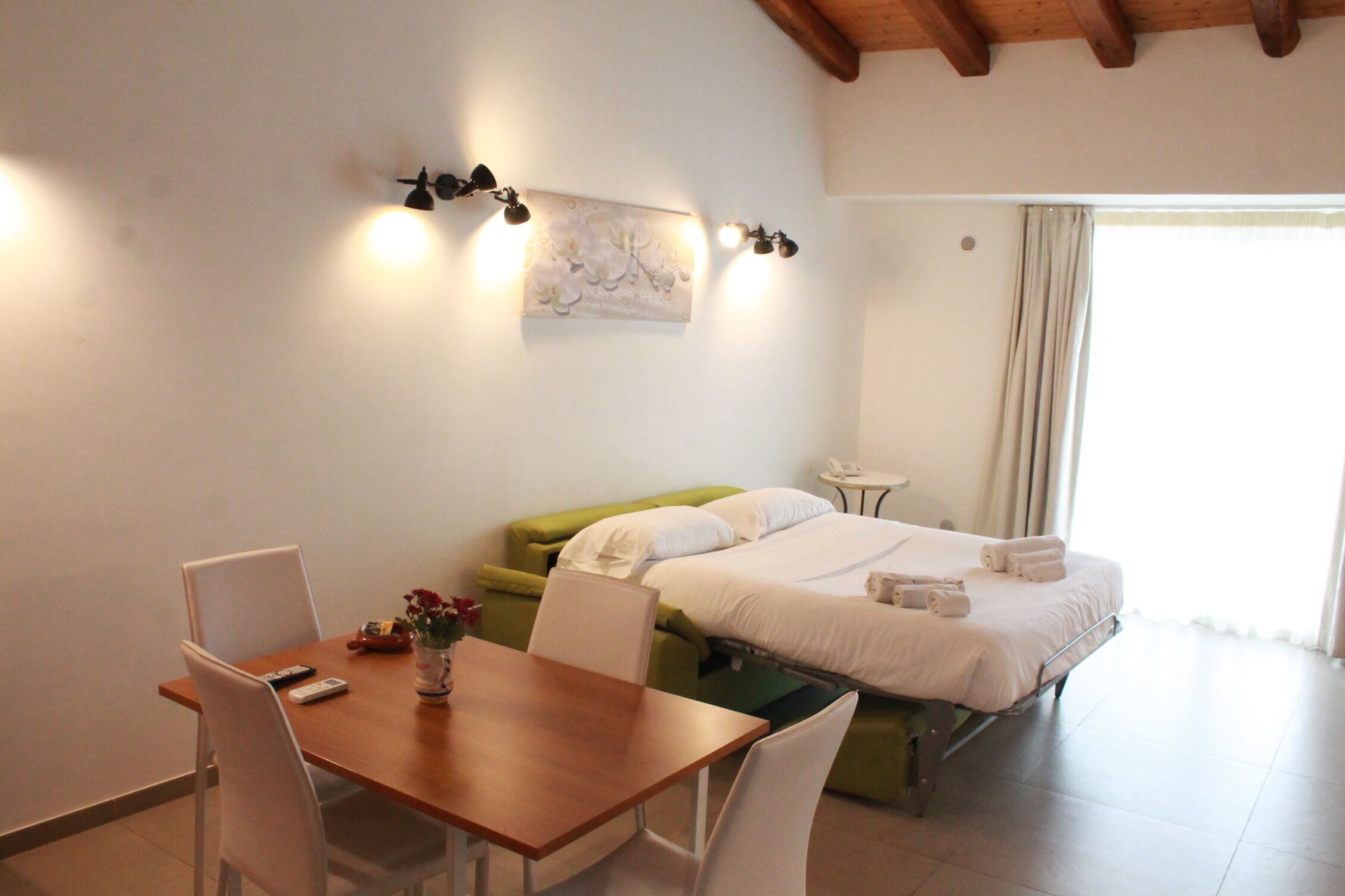 A hotel room with a wooden table, two white chairs, a green sofa, a bed with white linens, and a wall-mounted light fixture.