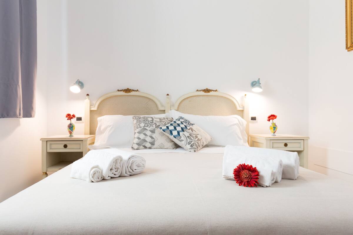 Un lit bien fait, recouvert d’une couette blanche, avec des oreillers et une fleur rouge. Il y a deux tables de chevet, chacune avec une lampe et un vase de fleurs.