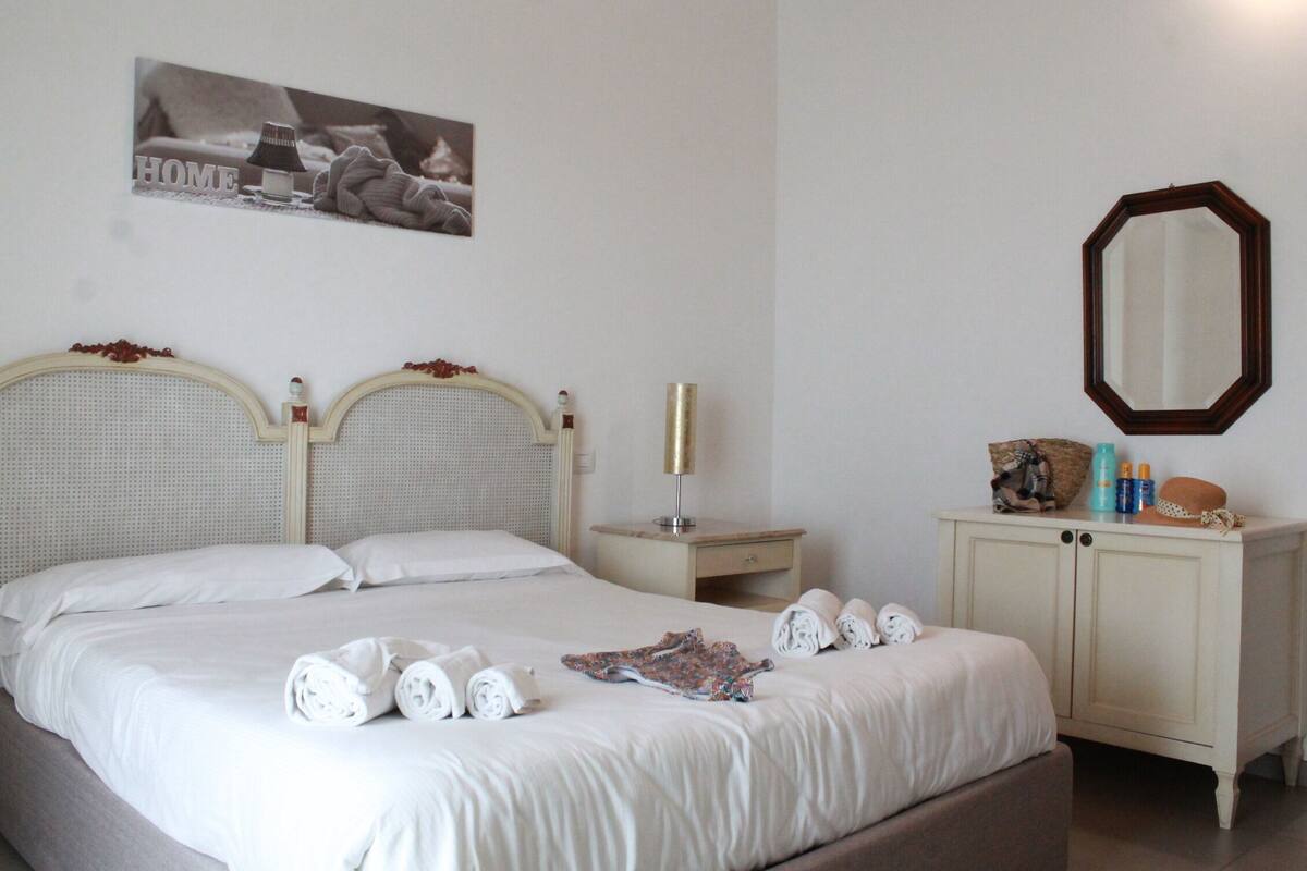 Un lit bien fait, avec des serviettes pliées, une table de chevet avec une lampe et une commode contenant des articles de toilette.