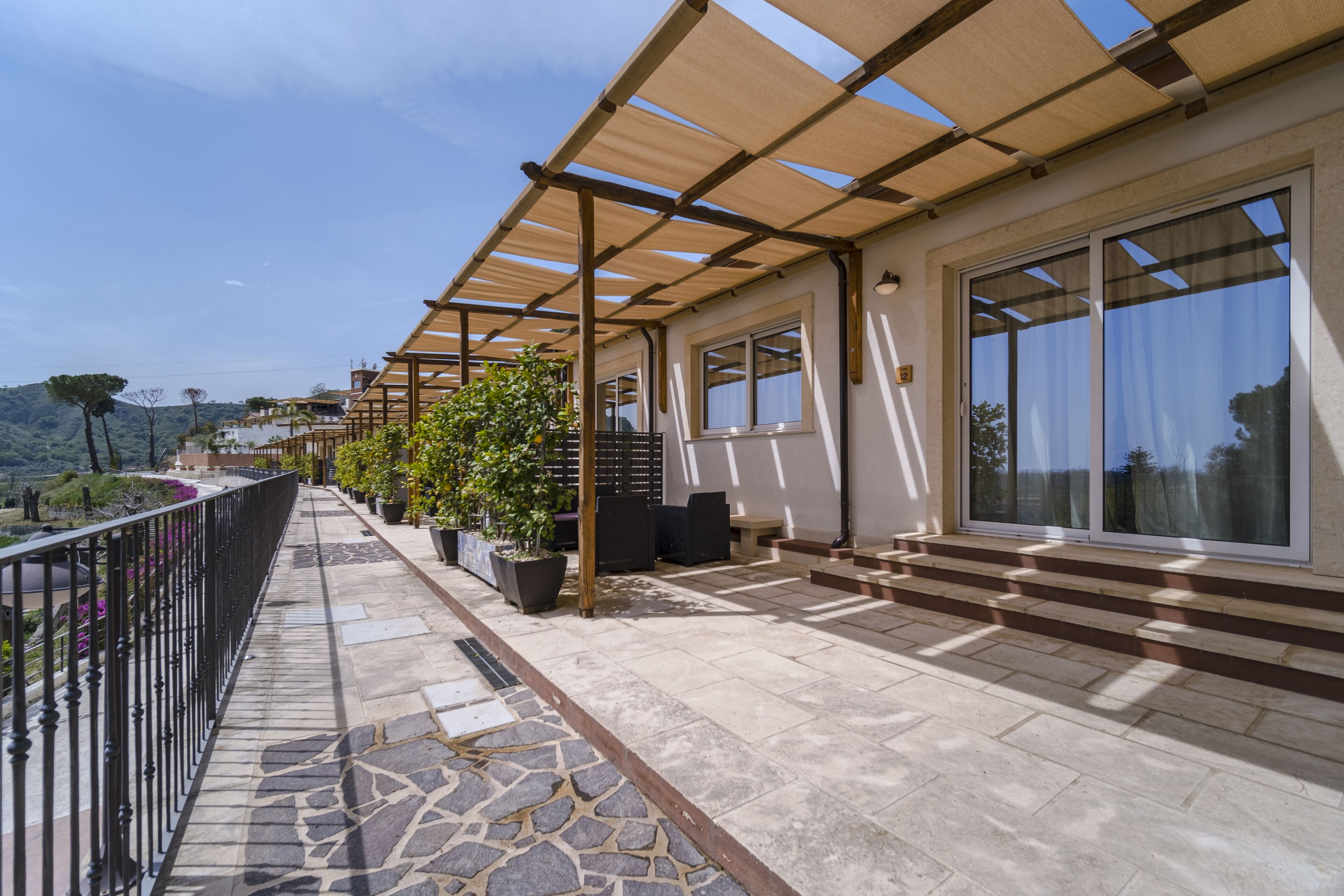 A balcony with a stone floor, potted plants, and a pergola providing shade.