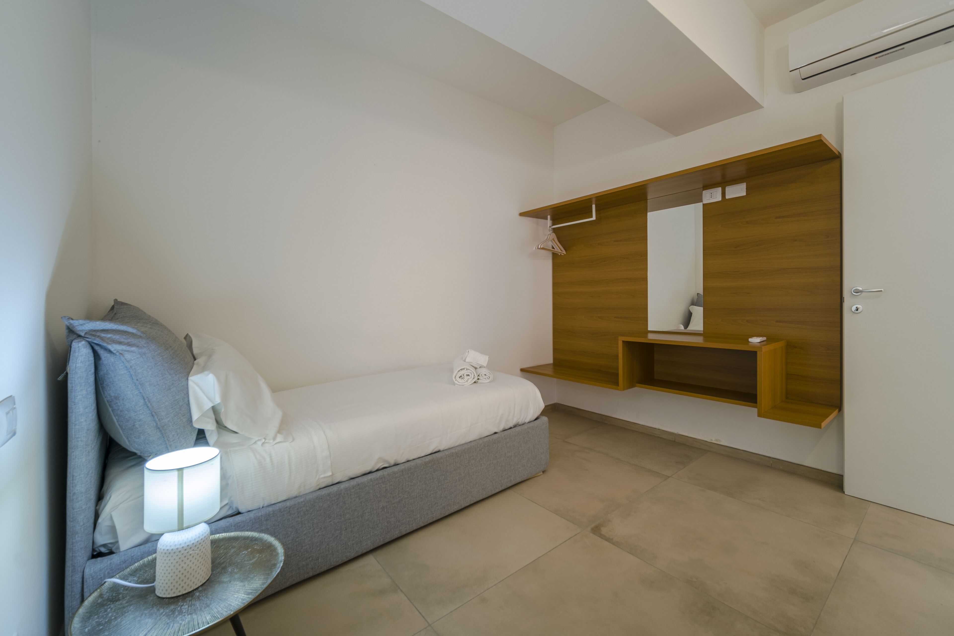 Ein modernes Schlafzimmer mit einem Bett, einem Nachttisch mit Lampe, einem Holzregal mit Spiegel und einer Tür.
