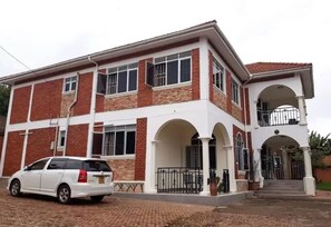 A two-story house with a white facade and red brick accents, featuring a covered porch and a car parked in front. - Emra Hotel (Entebbe)