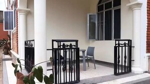 Un edificio con fachada blanca, una reja de metal negro, una mesa con una silla y una ventana con barrotes.