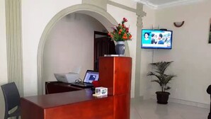 Un área de recepción con un escritorio de madera, una computadora portátil, un florero con flores, un televisor y una planta en maceta.