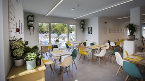 Un interno ristorante moderno con diversi tavoli e sedie, ampie finestre che offrono vista all'esterno e numerose piante in vaso.