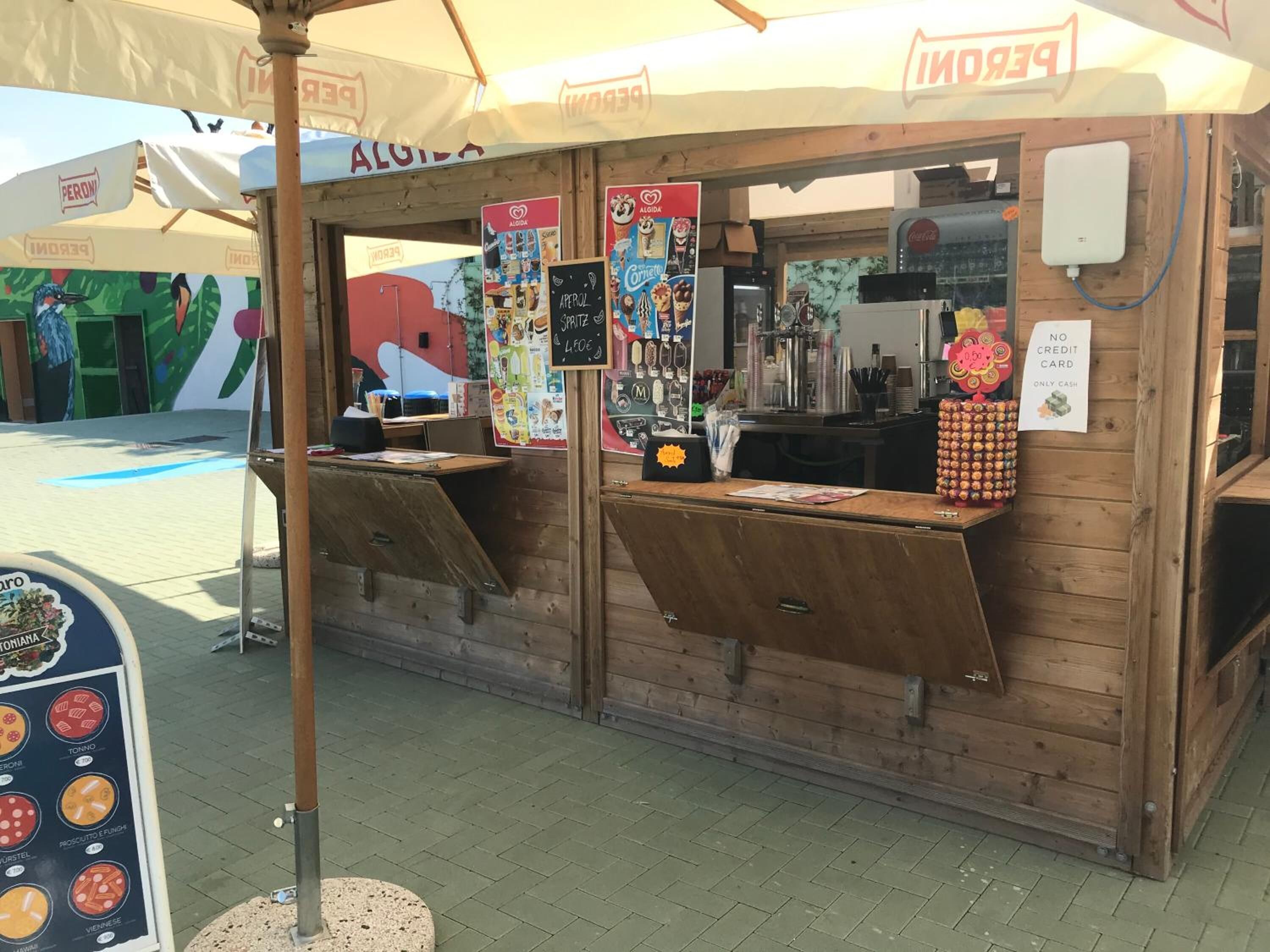 A food kiosk with a menu board, a Coca-Cola refrigerator, and a 'NO CREDIT CARD' sign.