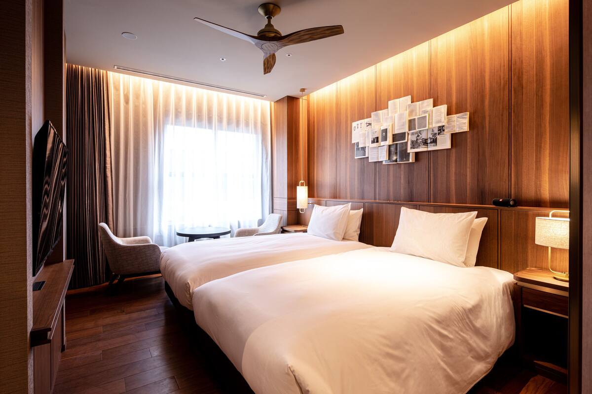 A hotel room with two beds, a desk, a chair, a ceiling fan, and a wall with photos.