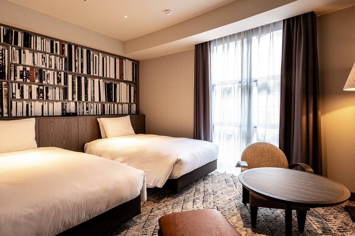 A hotel room with two beds, a bookshelf, a round table, and a chair.
