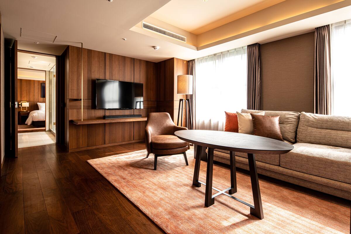 A modern hotel room with a wooden floor, a sofa, a table, and a flat-screen TV mounted on the wall.