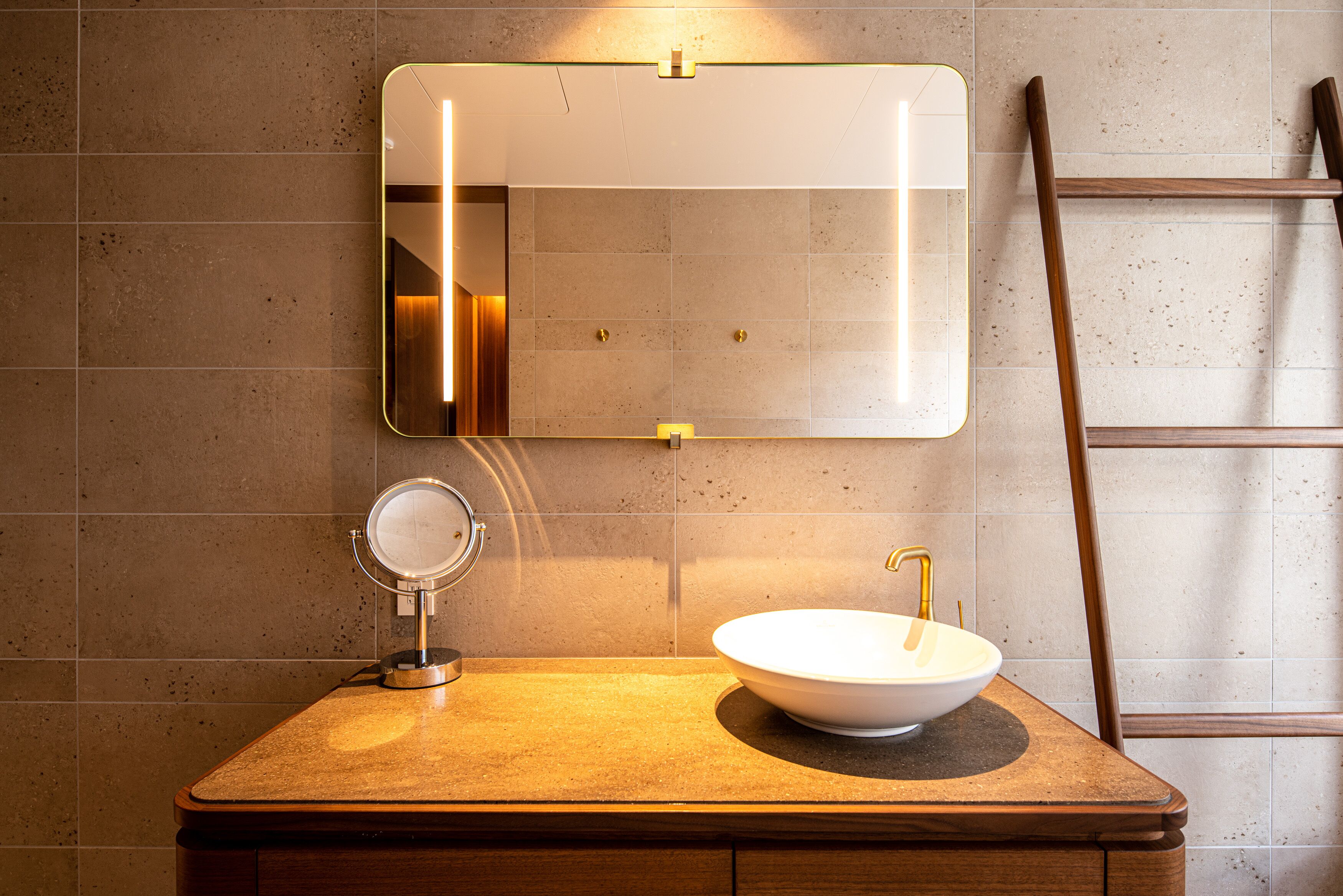 Un baño con tocador de madera, un espejo redondo, un lavamanos blanco con grifo dorado e iluminación integrada en el espejo.