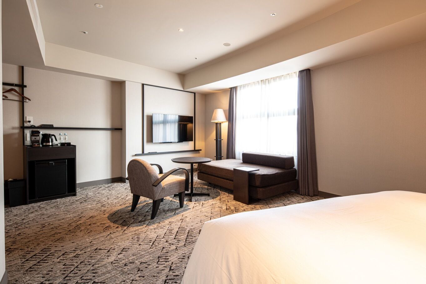 A hotel room with a bed, a desk with a lamp, a chair, a sofa, and a TV mounted on the wall.